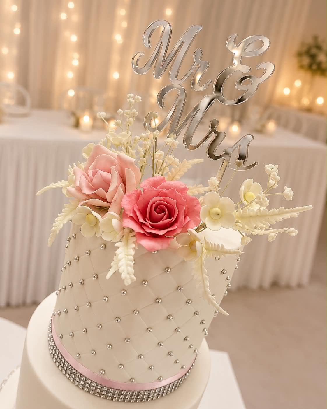 Close up of white quilted fondant wedding cake tier with silver pearl dot grid, pink and coral sugar roses, white gypsophila and fern sprigs and a silver Mr & Mrs acrylic topper from a different angle